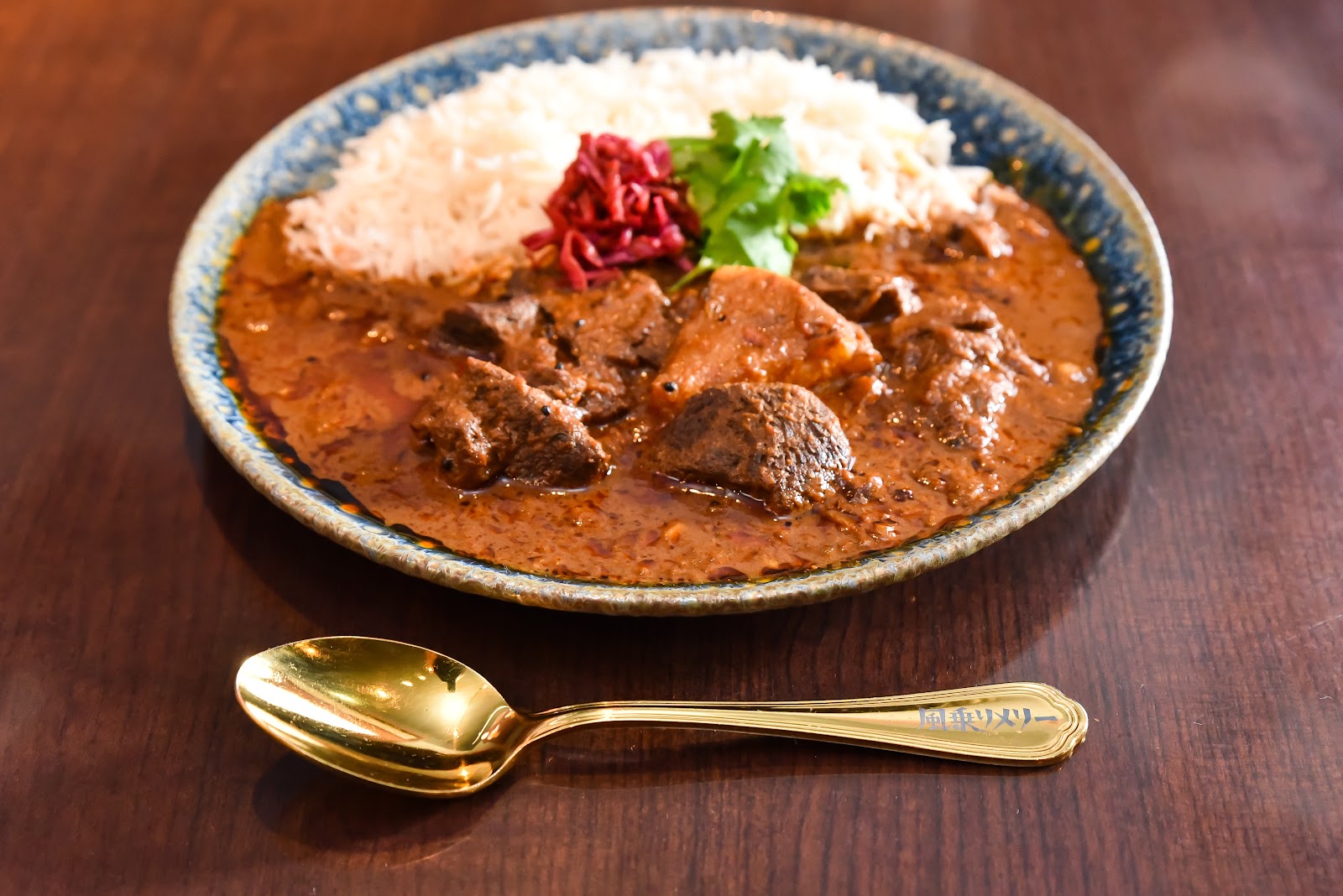 青い縁の陶皿に、黒っぽいカレーとバスマティライス。パクチーと赤い酢漬けの玉ねぎが添えてある。手前の木のテーブルに、柄に「風乗りメリー」と彫られた金色のスプーンが置かれている。