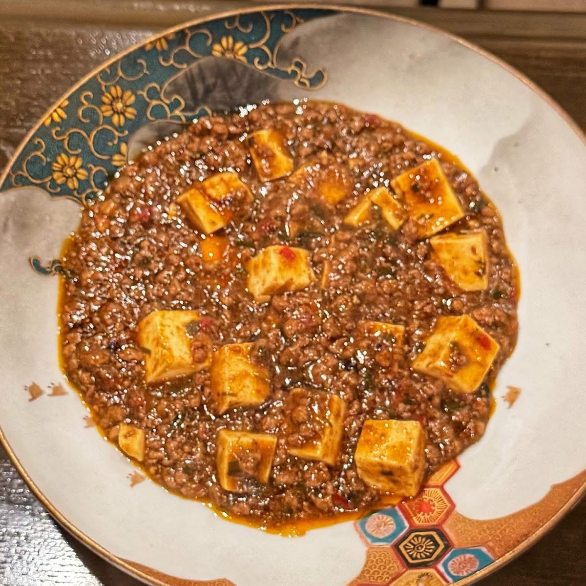真上から見下ろした麻婆豆腐。角切りの豆腐とひき肉のあんが皿いっぱいに盛られ、縁には金色の菊唐草と青緑の帯、右下に色絵の六角つなぎの文様がある。