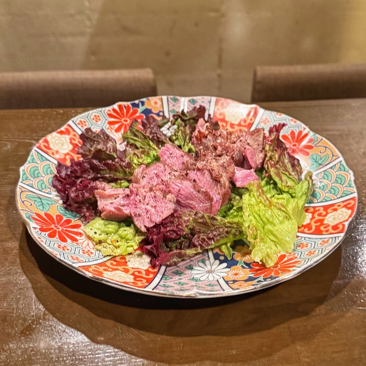 朱色と藍の色絵の大皿に、薄く切った赤身の肉が並び、赤と緑のリーフレタスが添えられている。皿の縁は菊の花の文様。奥は板張りのテーブルとソファ席。